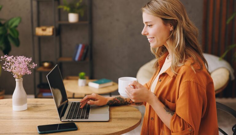 Side view young IT woman wearing orange shirt hold use laptop pc computer drink coffee