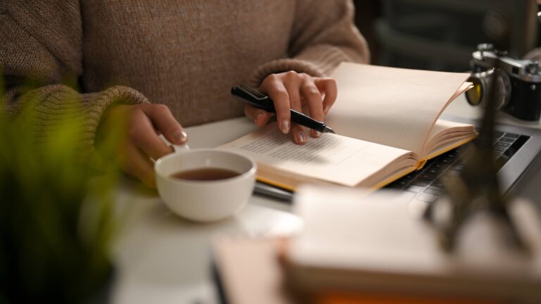 A female reading a book or writing a diary and sipping hot tea in cozy home workspace