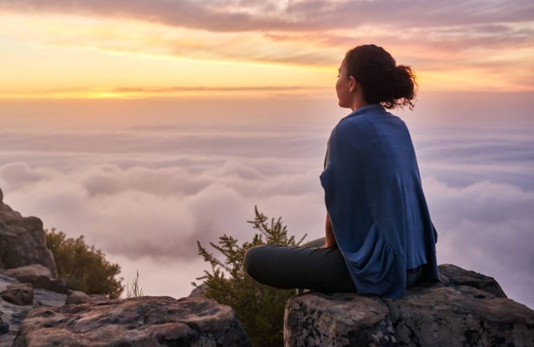 Rear view of a young woman sitting on a mountain top peacefully gazing at low-lying morning clouds